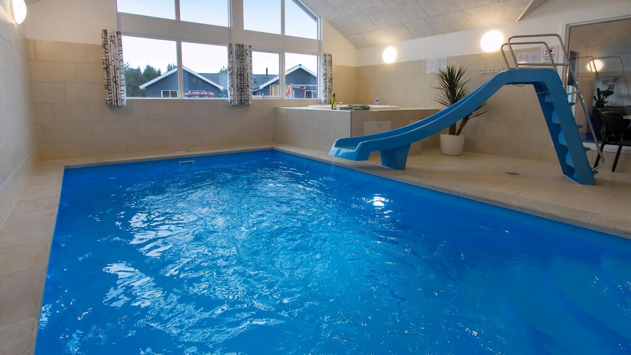 Een binnenzwembad met een blauwe glijbaan, grote ramen en uitzicht op huizen.