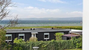 Un edificio moderno de color negro con techo plano, rodeado de vegetación, con vistas a un cuerpo de agua y montañas distantes.