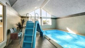 Une piscine intérieure avec un toboggan, un espace baigné de lumière avec des chaises, et une vue sur l’extérieur.