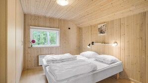 Une pièce aux murs lambrés, comprenant un lit, une fenêtre et une étagère fixée au mur où sont rangés des verres.