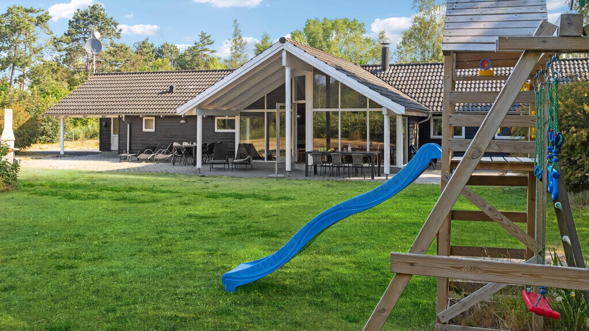 A playground with a blue slide and climbing area, a covered seating area with tables and chairs, and a building with a tiled roof.