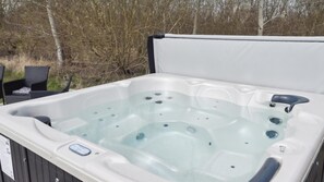 Un jacuzzi con revestimiento blanco y detalles negros, rodeado de mobiliario exterior y árboles.