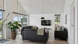 A spacious living room with a large black sofa, a smaller sofa, a TV mounted on the wall, and a large window offering a view of the outdoors. - VillaVilla 614 - Hasmark Strand, Fyn og øer (Otterup)