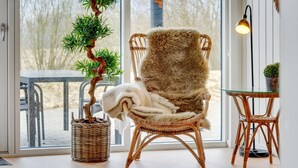 Une chaise en osier agrémentée d’un plaid en fourrure, une petite table sur laquelle est posée une plante en pot, et une grande fenêtre donnant sur du mobilier d’extérieur.