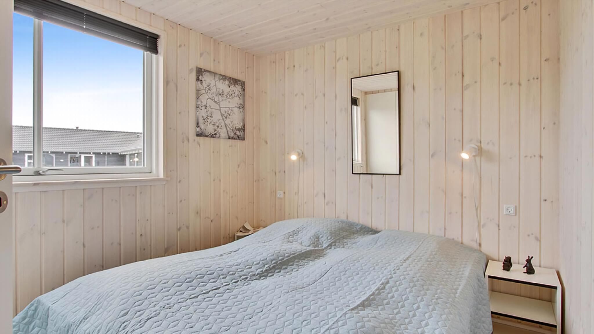 A bedroom with a bed, a window, a wall-mounted mirror, and a small bedside table with two figurines.