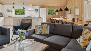 Ein modernes Wohnzimmer mit einem grauen Sofa, einer Essecke mit einem Holztisch sowie einer Küche mit weißen Schränken und einem Fenster.
