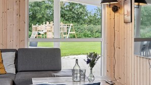 Ein Zimmer mit einer grauen Couch, einem Tisch mit einer Vase und Gläsern sowie einem Blick auf eine Holzterrasse und Grünflächen.