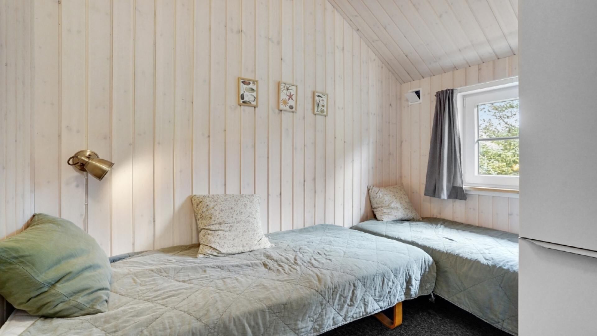 A small, single-room cabin with two beds, a window with curtains, and a wall-mounted lamp.
