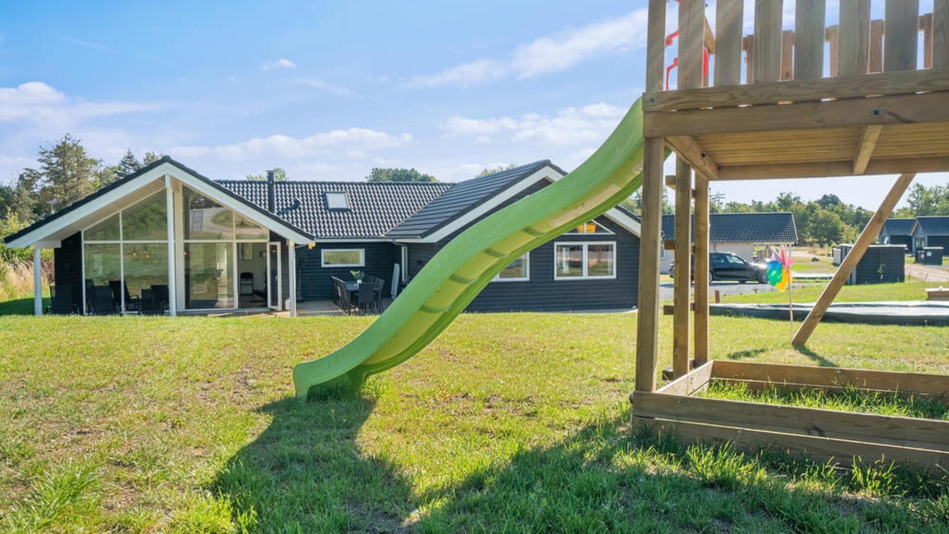 A modern house with a large glass front, a wooden play structure with a slide, and a grassy yard.
