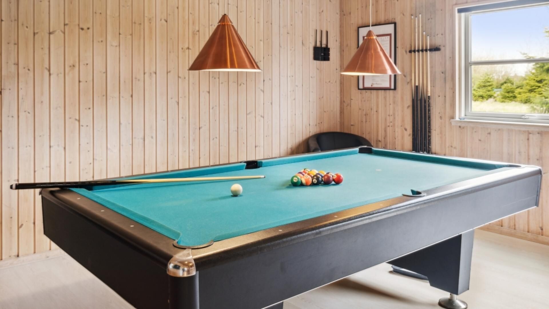 A pool table with balls, two pendant lights, a framed certificate on the wall, and a window with a view of trees.