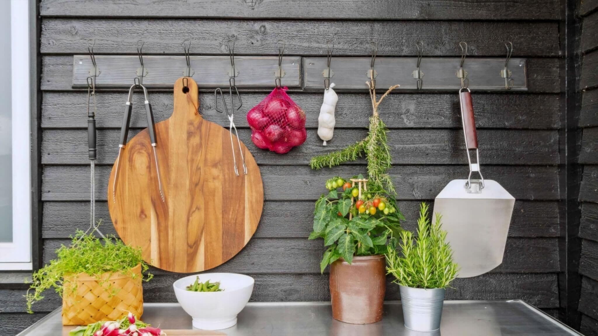 Un plan de travail de cuisine avec une planche à découper en bois, un sac en filet rouge, un oignon blanc, une branche d’herbes aromatiques, une spatule en métal et plusieurs plantes en pot.