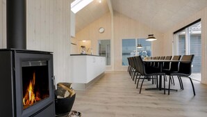 A modern kitchen with a wood-burning stove, a dining area with a long table and chairs, and a large window providing natural light. - VillaVilla 392 - Faaborg, Fyn og øer (Faaborg)
