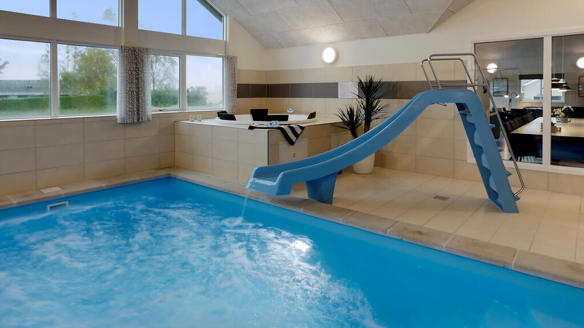An indoor swimming pool with a blue slide, a reception desk, and a view of the outdoors through large windows.