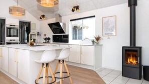 Et moderne køkken med en central ø, barstole, en sort brændeovn og et stort vindue.