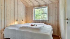 Une chambre avec un plafond en lambris de bois, un lit simple recouvert d’une courtepointe blanche, une petite table de chevet avec une lampe et une fenêtre donnant sur un paysage verdoyant.