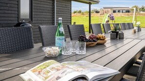 Eine Holzterrasse mit Gartenmöbeln, einer S.Pellegrini-Flasche, Gläsern, Schalen mit Trauben und Nüssen sowie einer Zeitschrift.