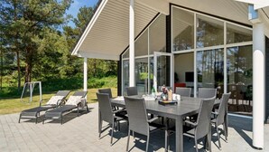 Une terrasse extérieure avec une table et des chaises, entourée de portes et de fenêtres en verre, donnant sur un jardin.