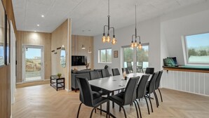 Uma sala de jantar moderna com uma mesa grande e cadeiras, piso de madeira e vista para o exterior através de amplas janelas.