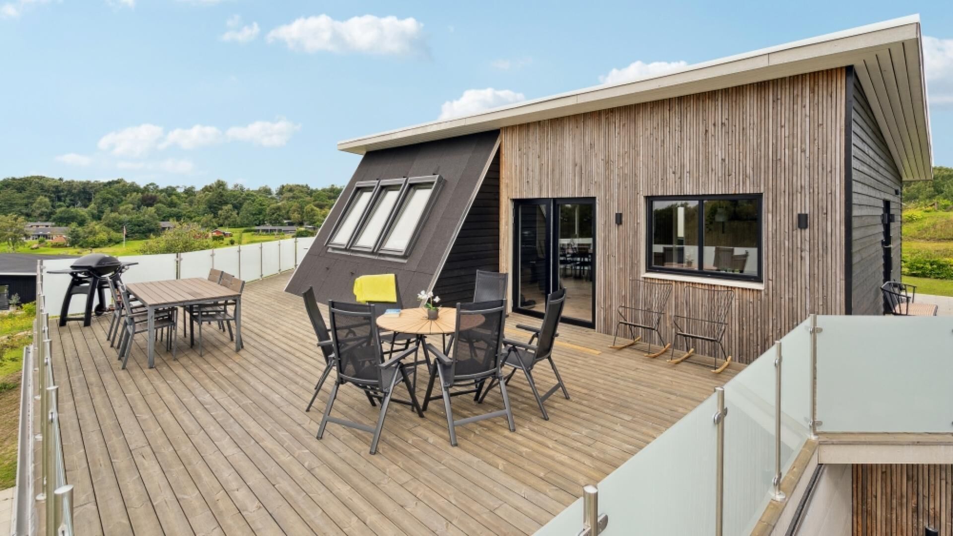 Une maison moderne dotée d’une terrasse en bois, d’un coin repas avec mobilier d’extérieur et d’une grande fenêtre donnant sur un paysage verdoyant.