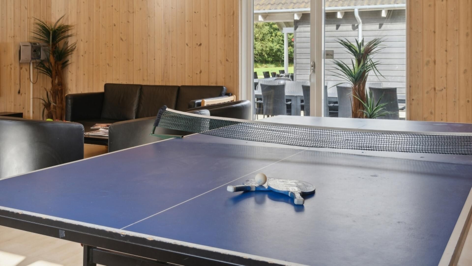 Een tafeltennistafel met net in een ruimte met houten wanden en uitzicht op tuinmeubilair op een terras.
