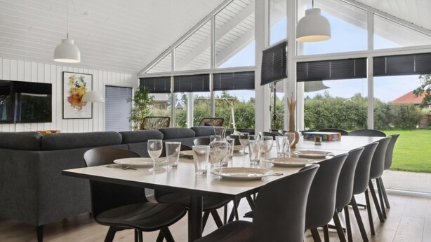 A modern dining area with a large table set for six, surrounded by dark chairs. There is a grey sofa and a dining table with a white top. The room has large windows with blinds, and a ceiling light fixture is visible. - VillaVilla 212 - Marielyst, Lolland/Falster/møn (Idestrup)