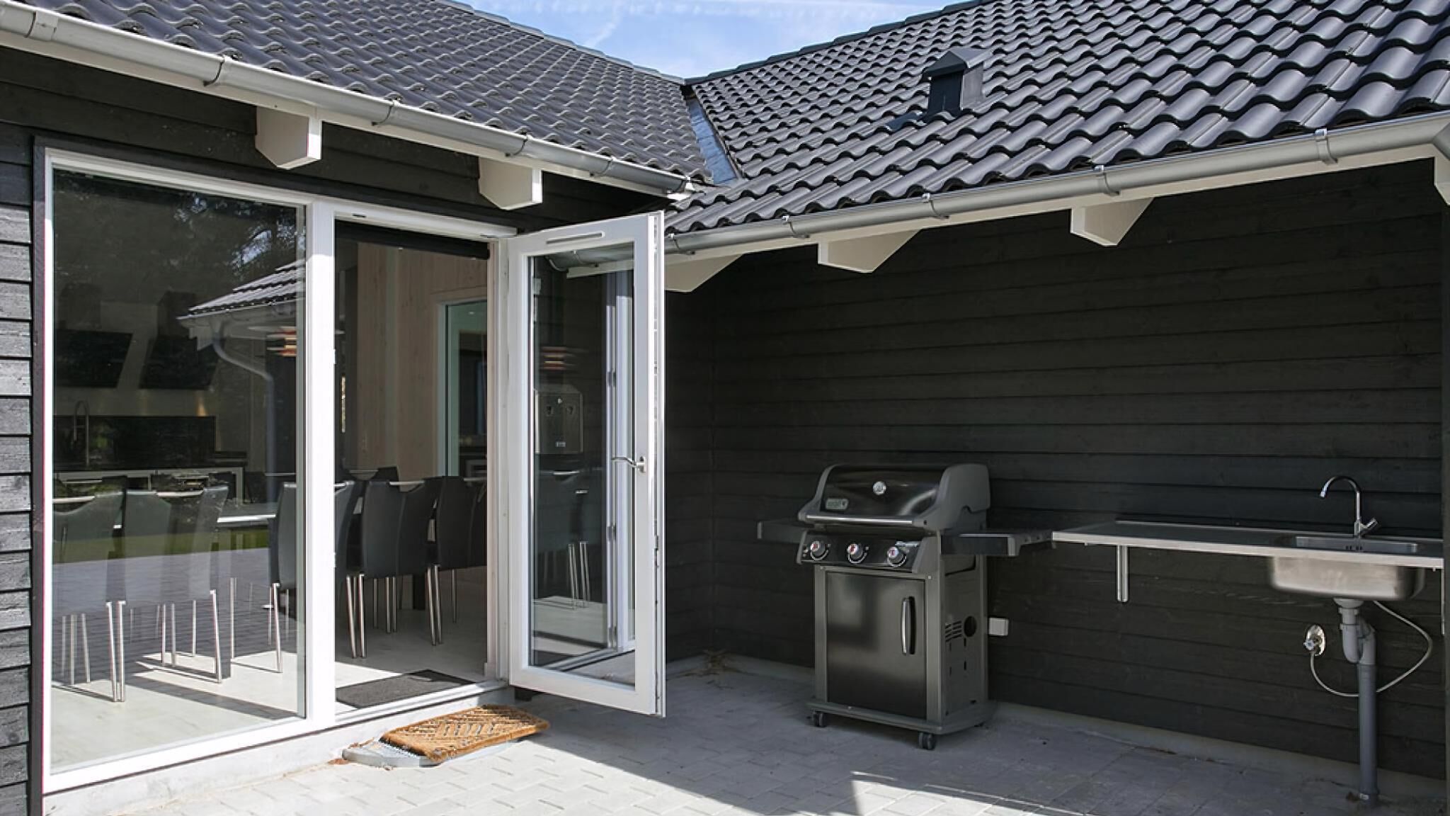 Ein Außenküchenbereich mit Grill, Spüle und einer Glastür zu einer Terrasse mit Sitzgelegenheiten.