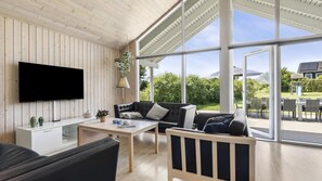 A modern living room with a flat-screen TV, a sofa, a coffee table, and a dining area with outdoor seating. - VillaVilla 274 - Tisvildeleje, Nordsjælland (Tisvildeleje)