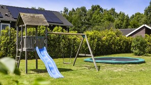 Puinen leikkialue, jossa on liukumäki ja keinut, trampoliini sekä taustalla aurinkopaneeleilla varustettu leikkimökki.