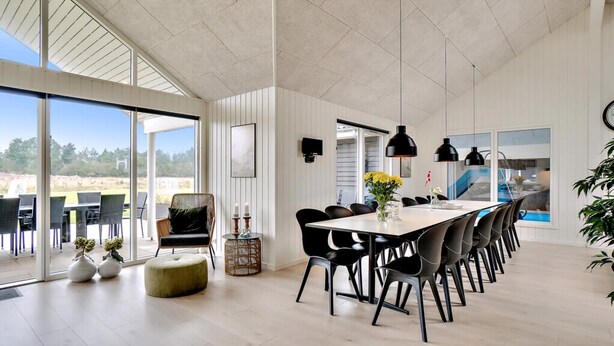 Uma ampla área de refeições com uma mesa comprida, cadeiras pretas, luminárias pendentes e vista para o exterior.