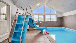 Piscina interna com um escorregador azul, uma janela e piso revestido de azulejos.