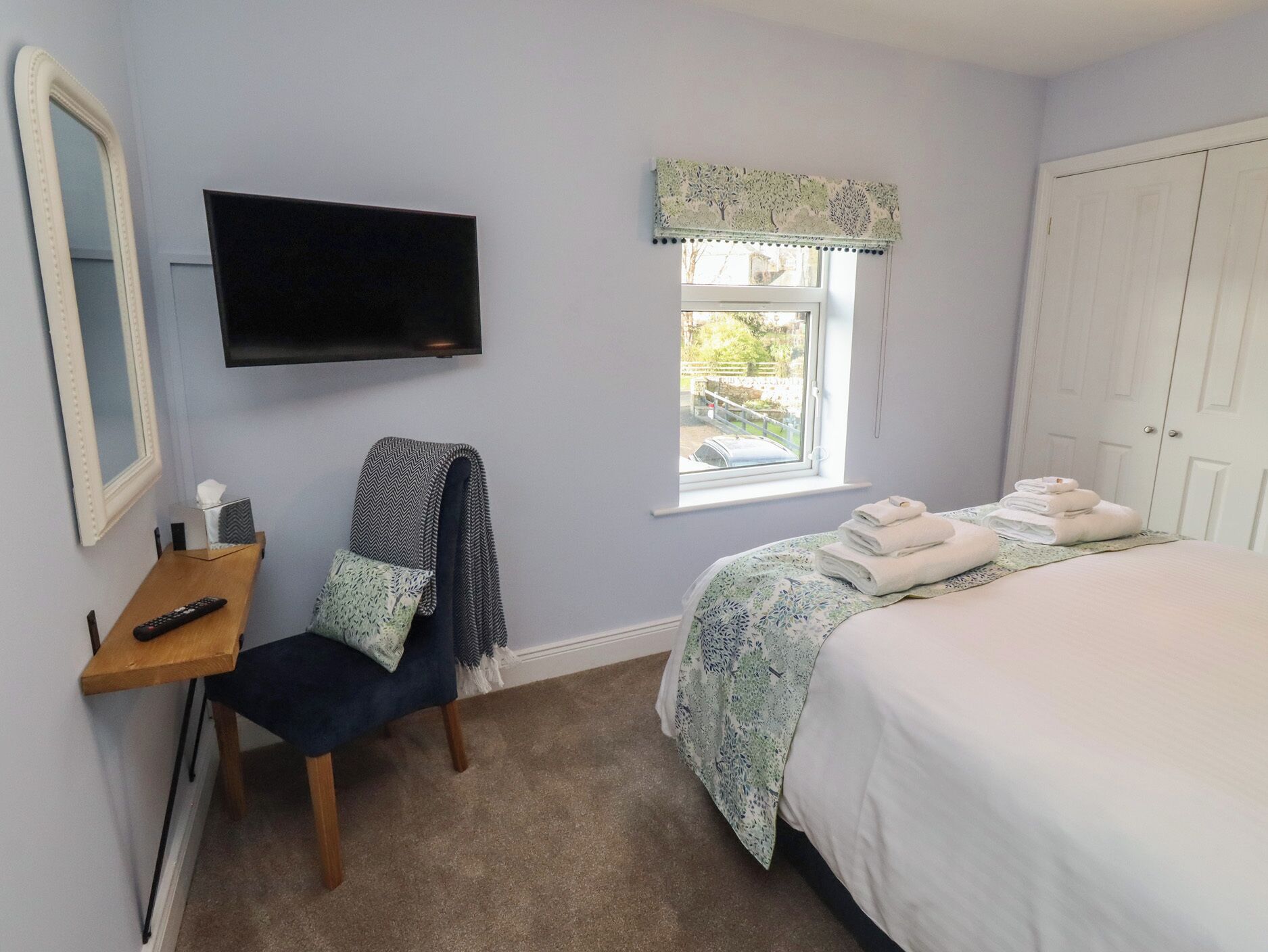 A bedroom with a bed, a TV mounted on the wall, a window with a view of the outside, and a door.