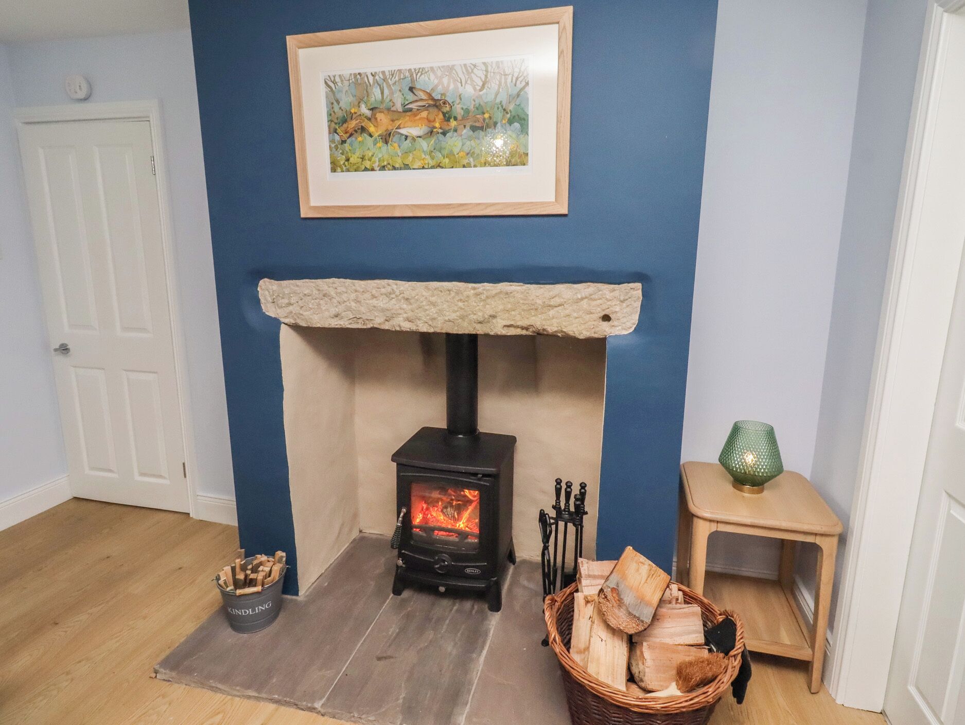 A room with a wood-burning stove, a framed painting on the wall, a door, and a small table with a green vase.