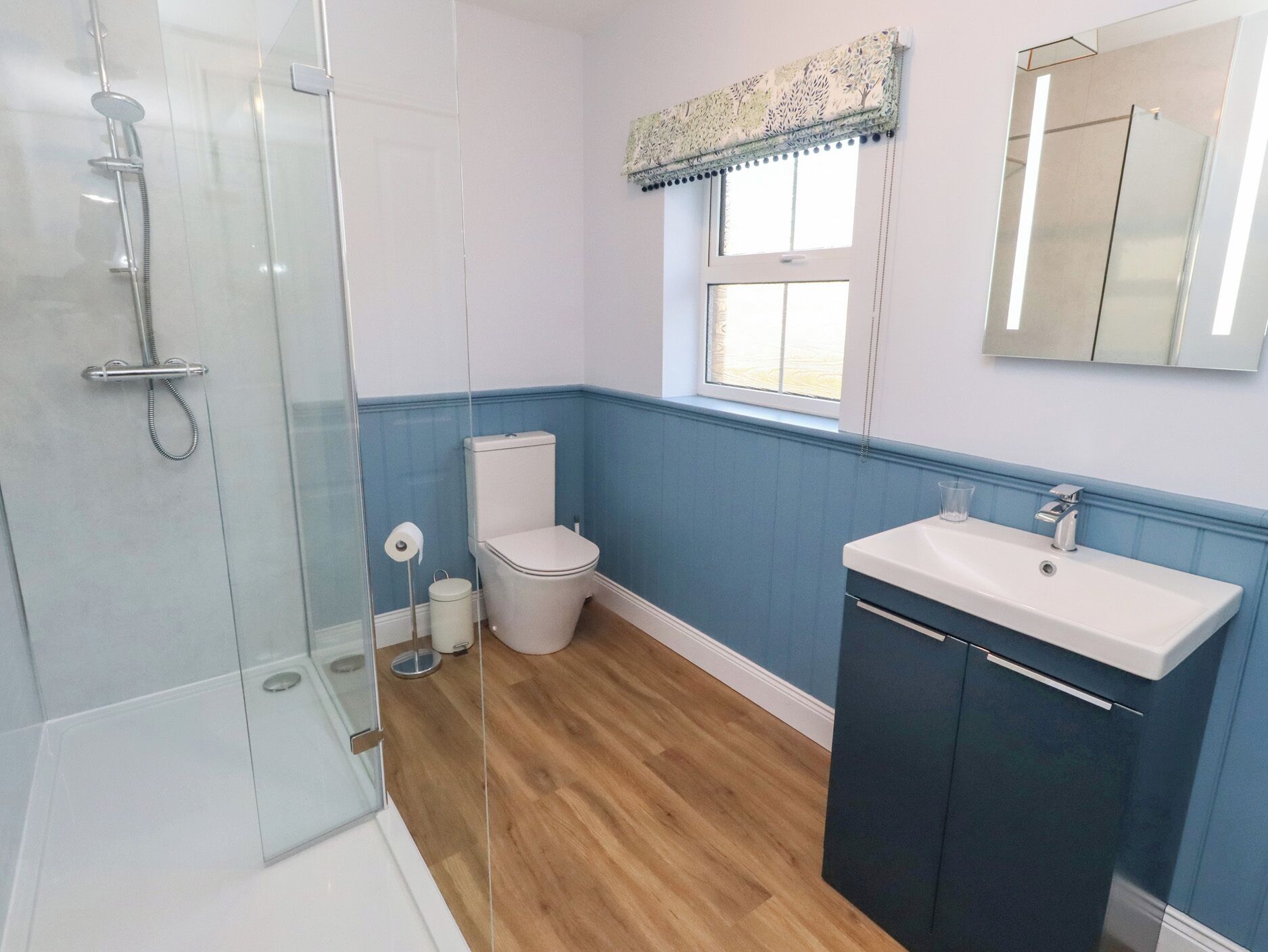 Un baño con cabina de ducha de vidrio, inodoro, lavamanos y ventana con un adornado cortinero.