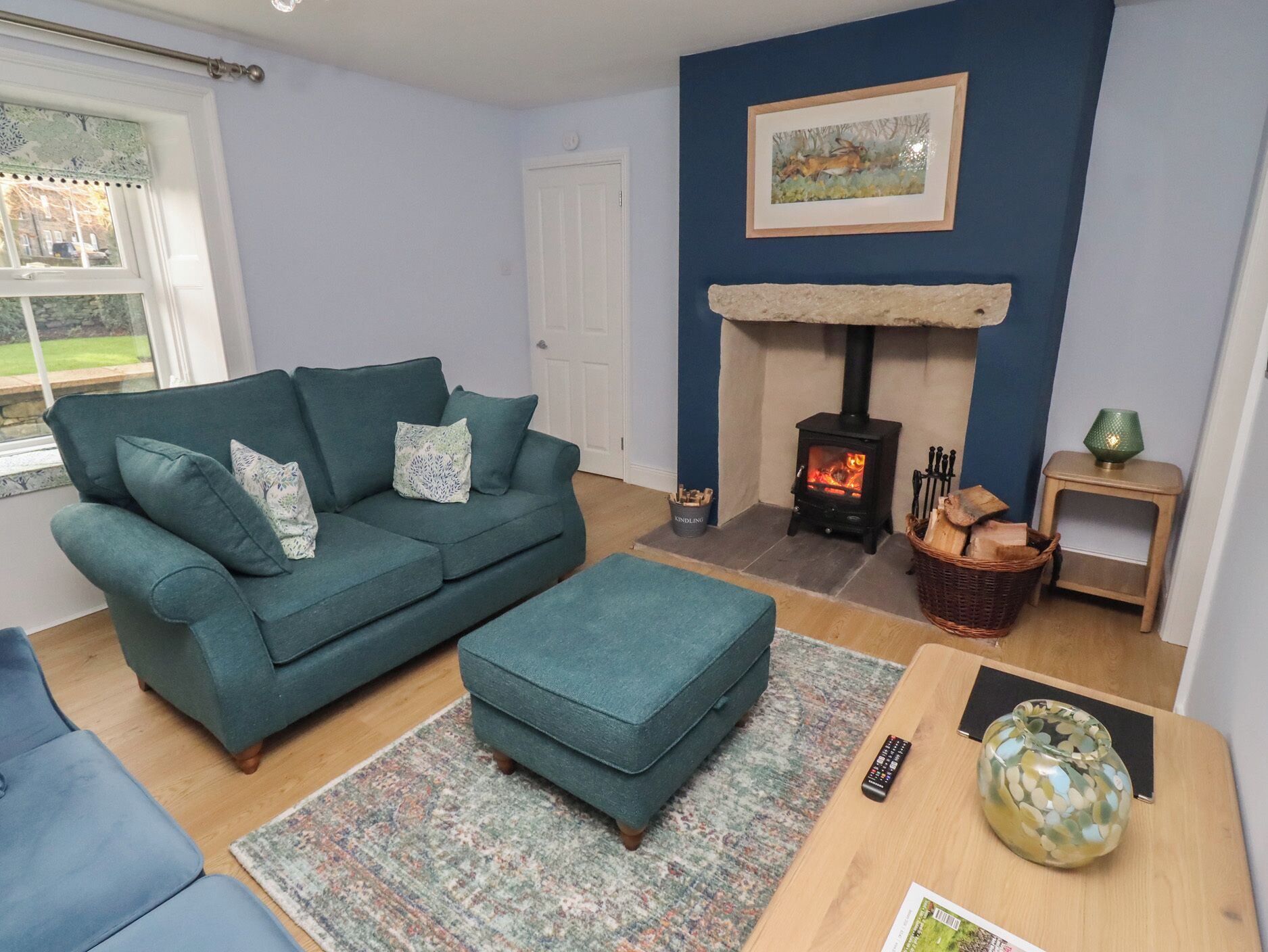 A cozy living room with a teal sofa, a matching ottoman, a fireplace, and a window with a view of the outdoors.