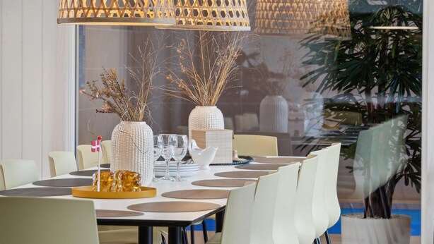 Un'area pranzo con tavolo imbandito per quattro persone, caratterizzata da un lampadario moderno, vasi bianchi con piante essiccate e un piccolo oggetto decorativo al centro del tavolo.