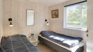 Et soveværelse med to senge, et lille natbord med en lampe, et billede på væggen og et vindue med udsigt til træer.