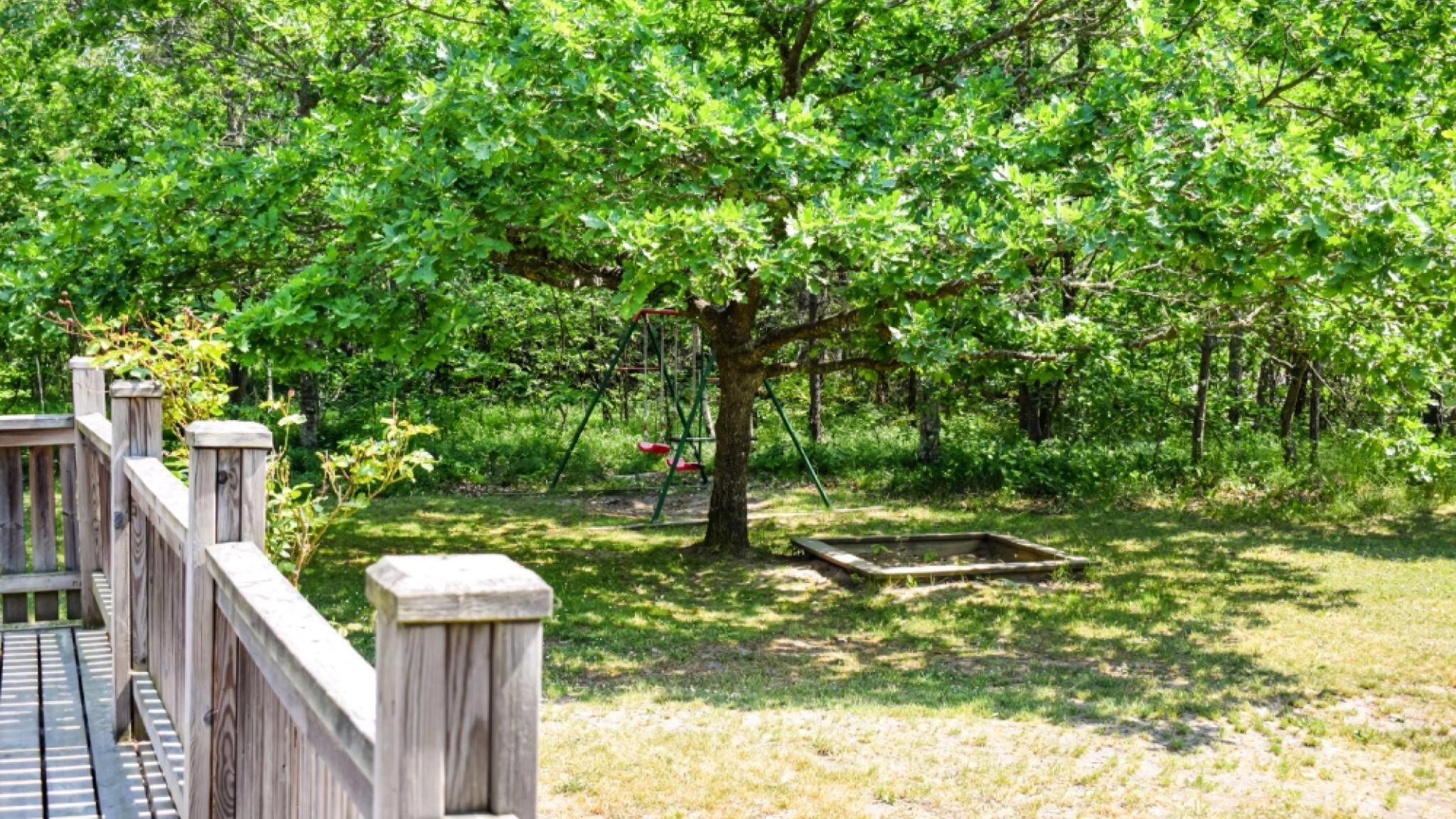 Una terraza de madera con un columpio y una pequeña construcción de madera en un área boscosa.