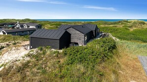 Ein modernes Haus mit dunklem Dach, auf einem grasbewachsenen Hügel mit Blick auf einen Strand.