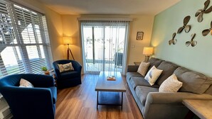 A living room with a blue armchair, a grey sofa, a wooden coffee table, and a floor-to-ceiling window with blinds.