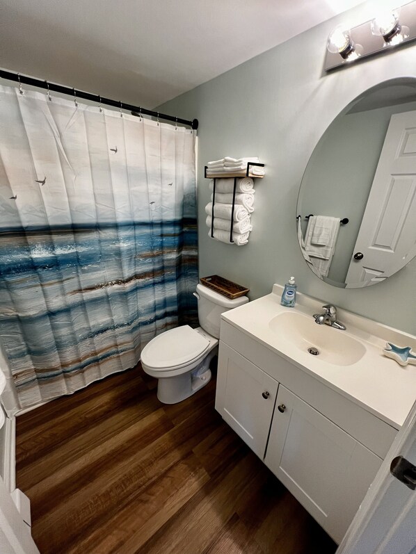 Un baño con suelo de madera, inodoro blanco, lavamanos blanco con espejo y cortina de ducha con estampado azul y blanco.