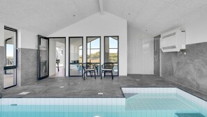 En moderne swimmingpool indendørs med udsigt til det fri, et siddeområde med stole, og et stort vindue.