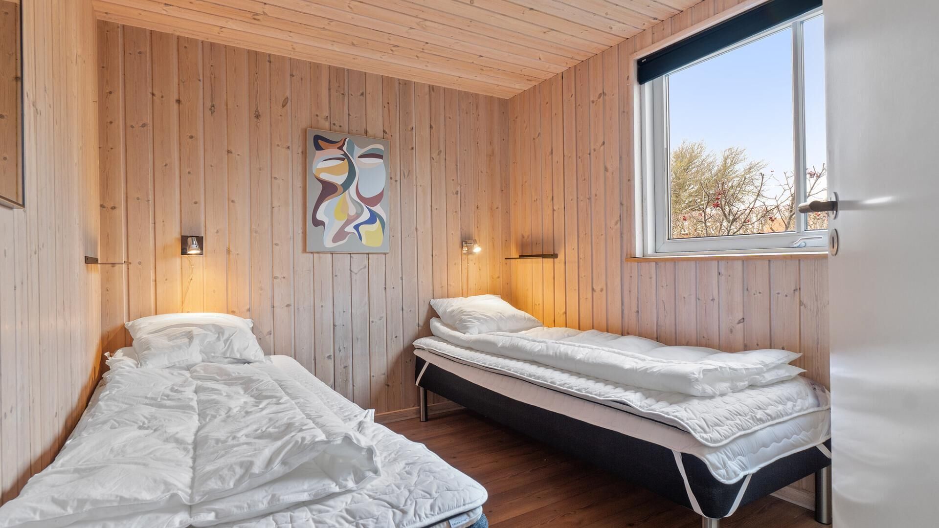 Una habitación con dos camas, paredes de madera, un ventanal grande y un cuadro abstracto colgado en la pared.