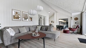 Een moderne woonkamer met een grijze modulaire bank, een ronde salontafel en een rode fauteuil. Er hangen ingelijste foto’s aan de muur, in de achtergrond is een eethoek en een trap die naar boven leidt.