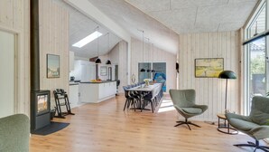 A modern kitchen with white cabinets, a dining area with a long table and chairs, and a cozy seating area with a green armchair and a floor lamp. - VillaVilla 494 - Thorup Strand, Nordjylland (Fjerritslev)