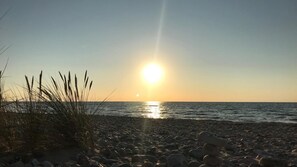 A beach with pebbles and grass, and the sun setting over the water.
