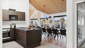 A modern kitchen with dark wood cabinetry, a central island, and a dining area with black chairs. - VillaVilla 584 - Hornbæk, Nordsjælland (Dronningmølle)