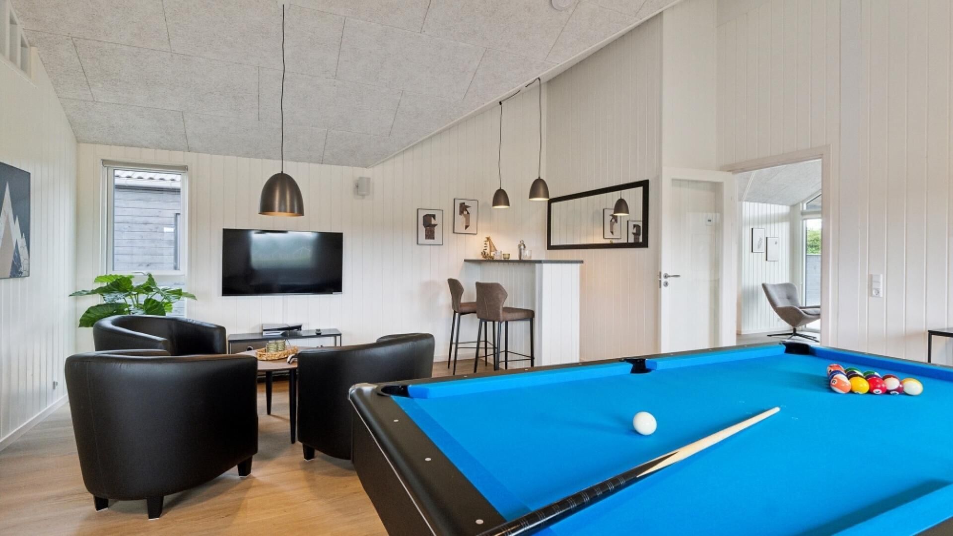 Et billardbord med blå filtdug, et poolstang og billardkugler. Der er et fjernsyn monteret på væggen, et barområde med barstole, og et siddeområde med stole.