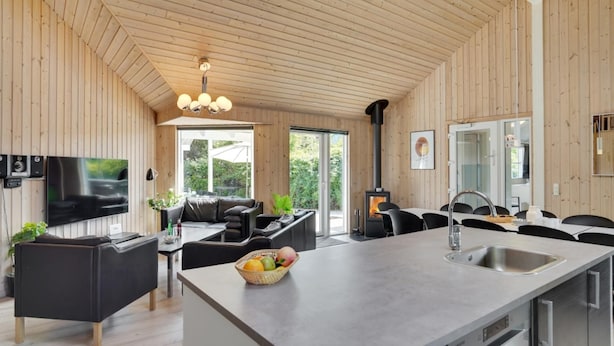 Ett modernt kök med en köksö i mitten, en matplats med svarta stolar, en platt-TV som är monterad på väggen och ett stort fönster som ger utsikt över omgivningen.