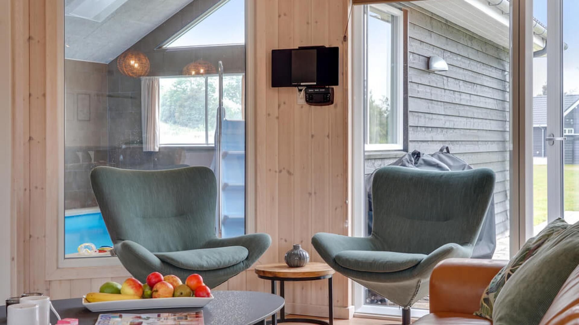 A modern room with two grey armchairs, a small round table with a vase, and a fruit bowl on a coffee table. A television is mounted on the wall, and there is a sliding glass door leading outside.