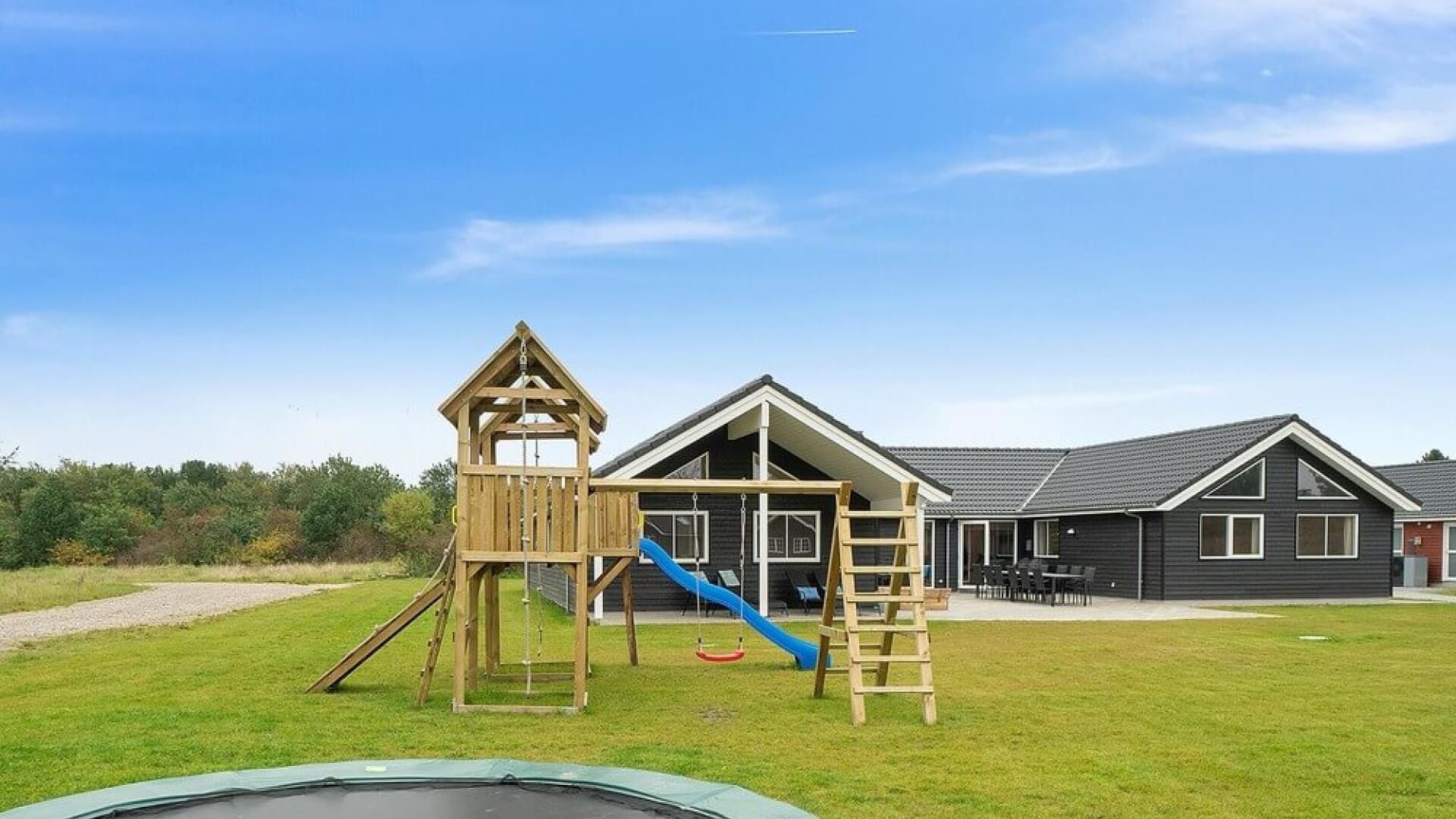 A playground with a wooden slide and climbing structures, a trampoline, and a building with a dark exterior and a sloped roof.